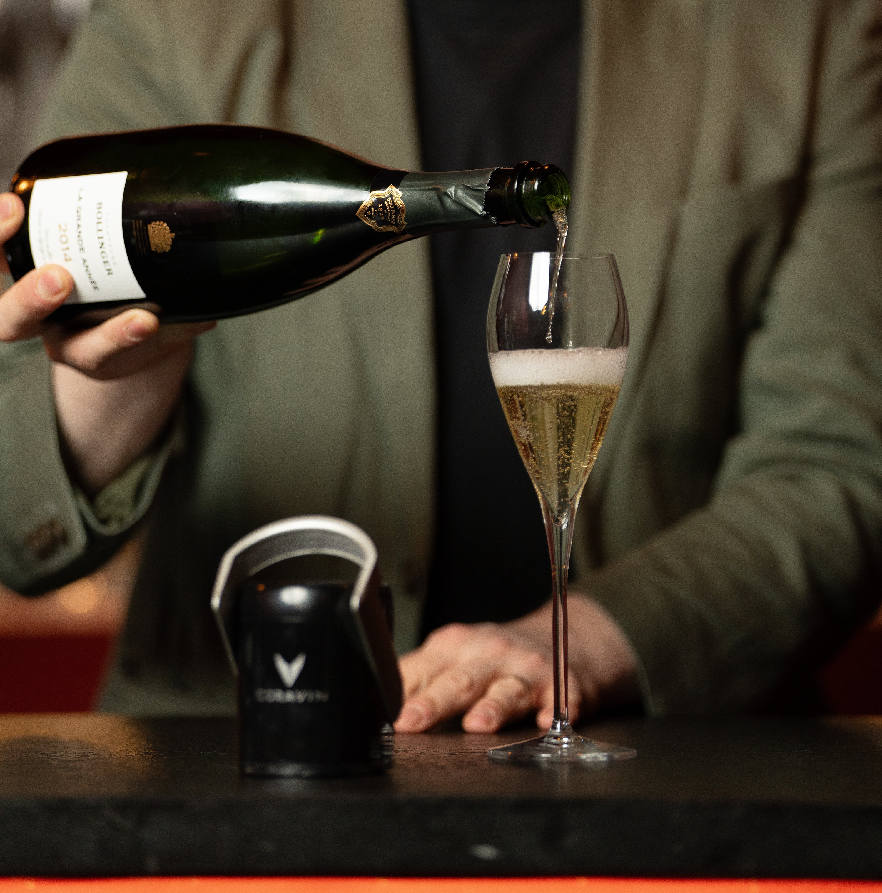 A bottle of Bollinger Champagne is poured into a flute beside the Coravin Sparkling® Stopper on a bar.