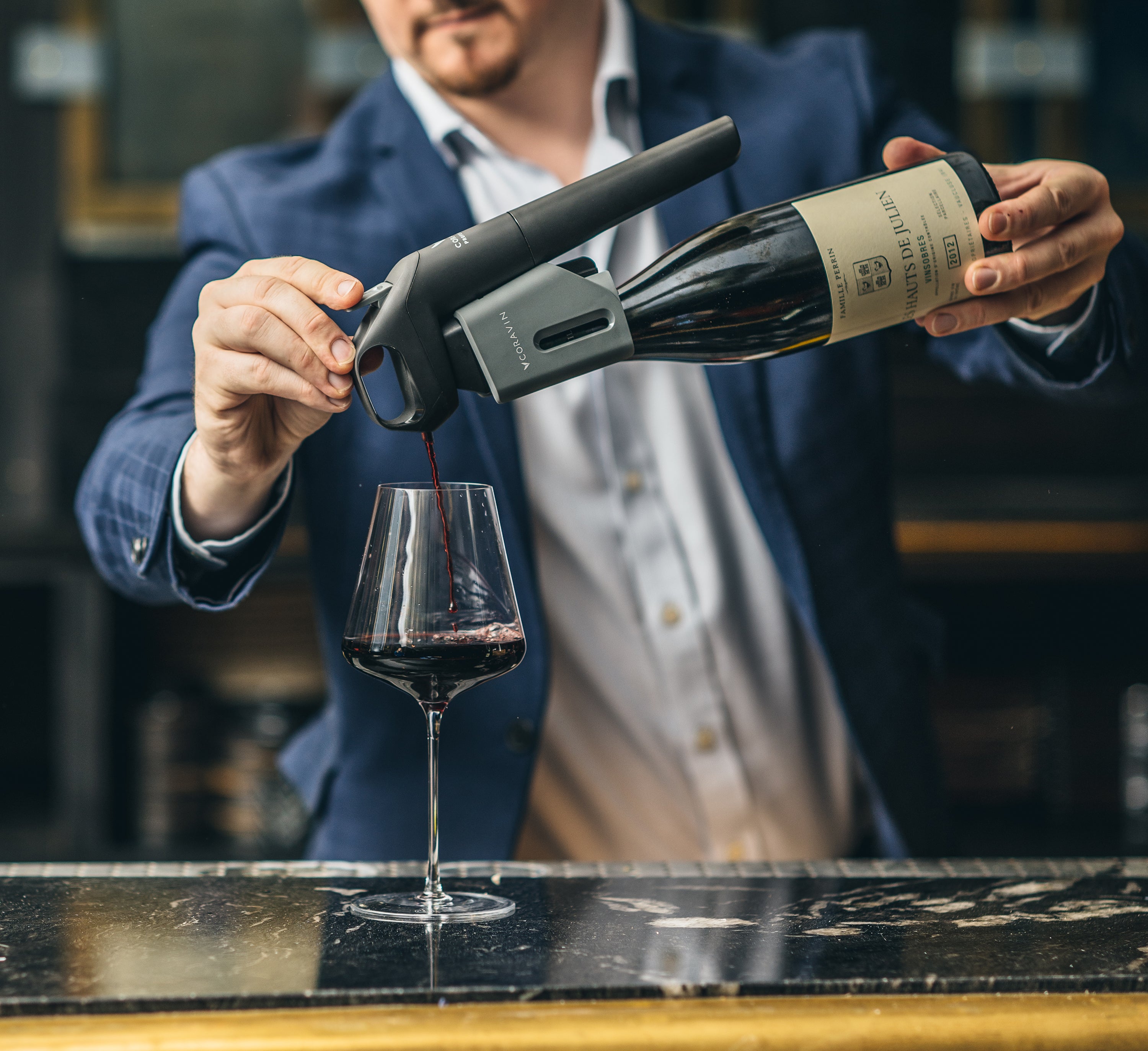 A man pours red wine using the Coravin Timeless system into a glass on a sleek black marble bar.
