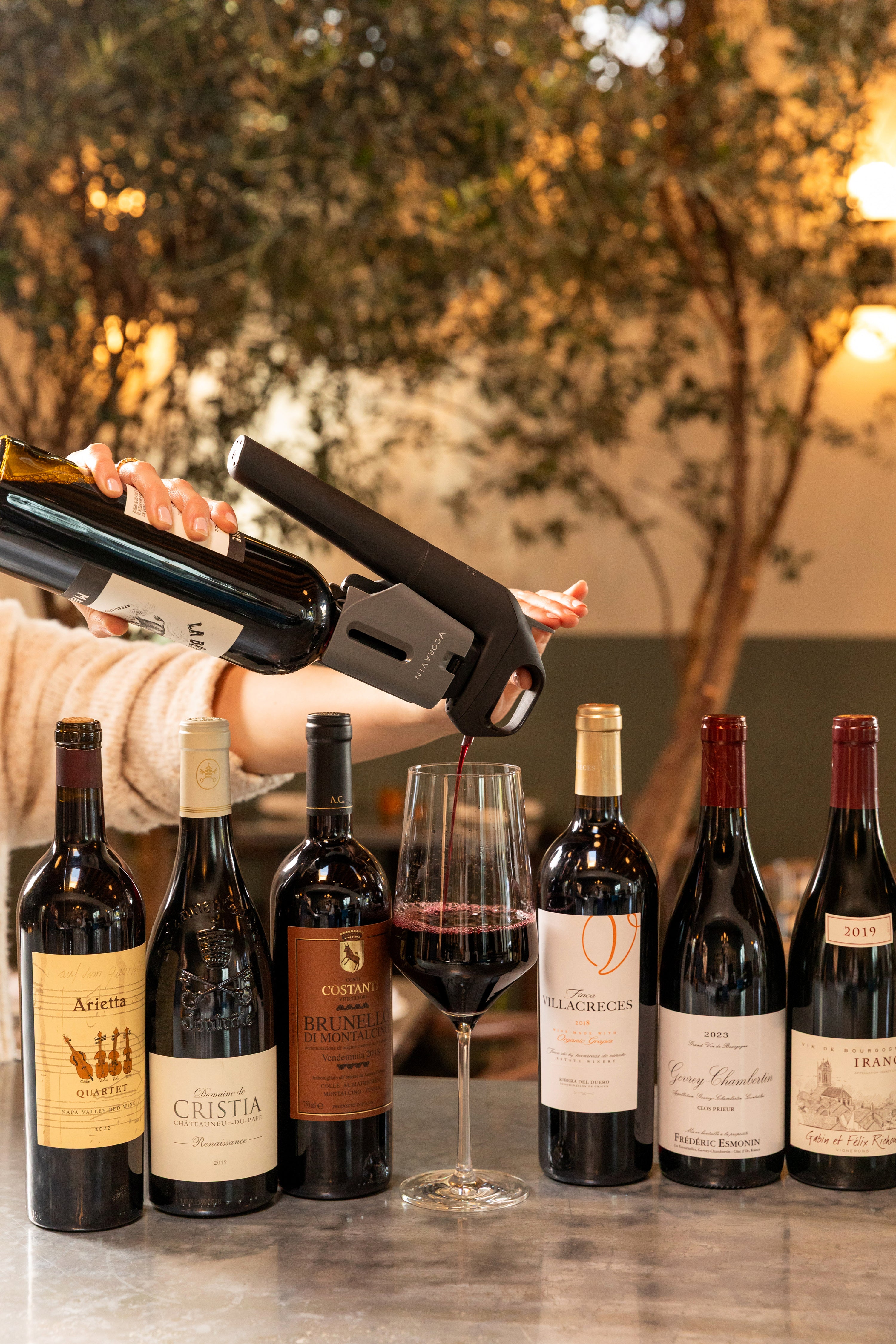 A Coravin Timeless system pours red wine into a glass, surrounded by premium still wine bottles.