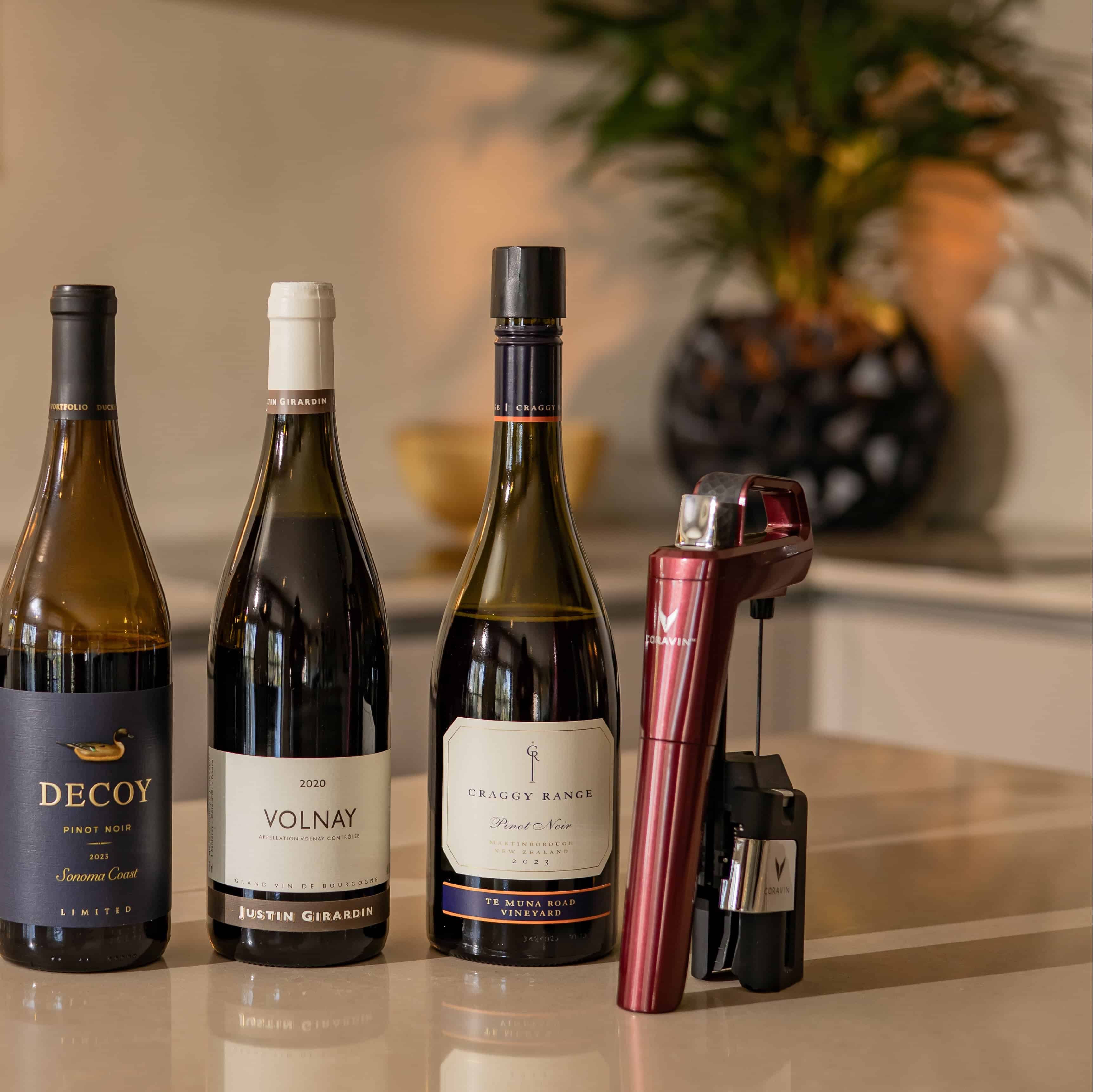Four wine bottles and a coravin red wine preservation system on a reflective surface with a blurred background