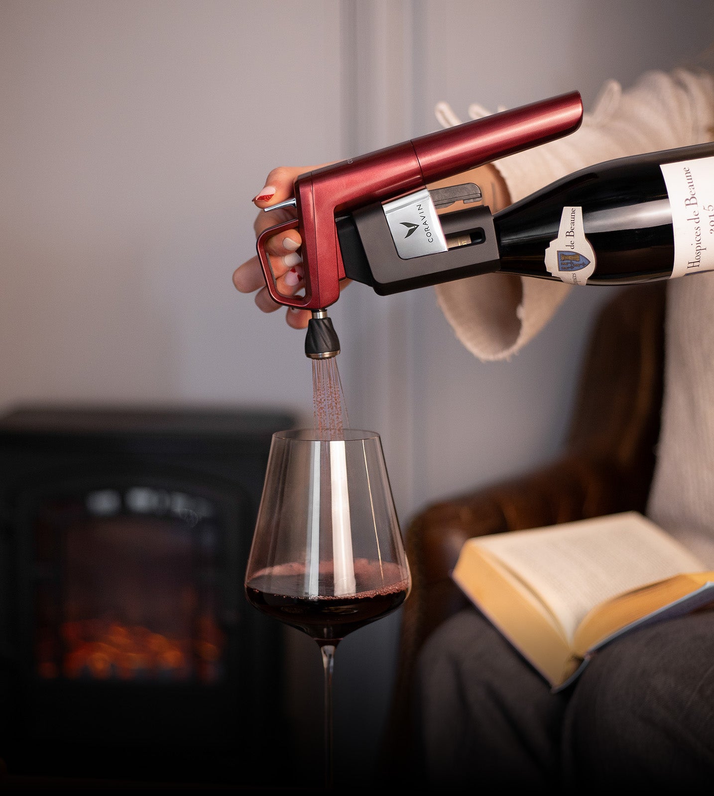 Person using a Coravin to pour red wine into a glass with a cozy indoor background.