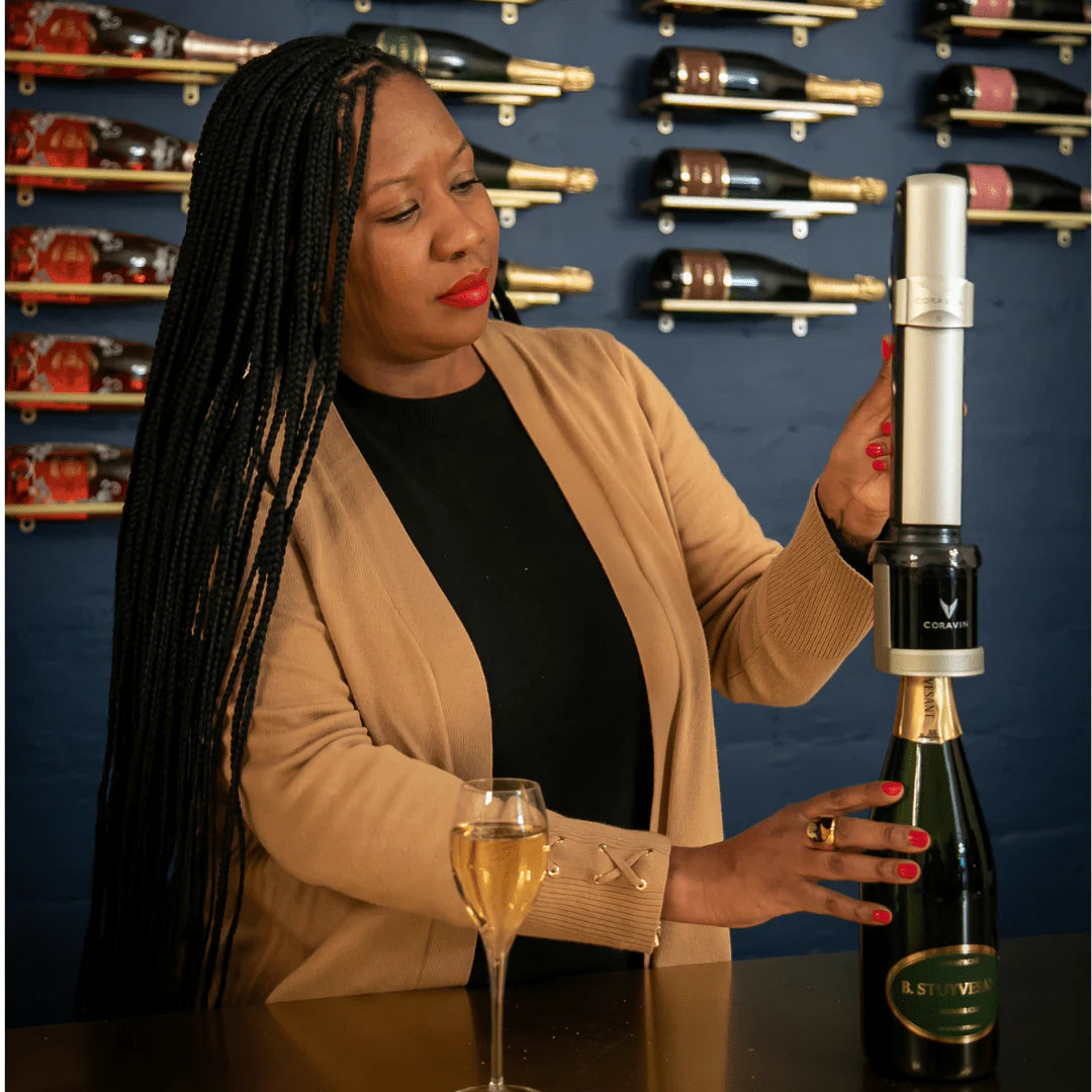 Woman holding a Coravin Sparkling and a wine bottle with a wine glass in front of a wall of wine bottles.