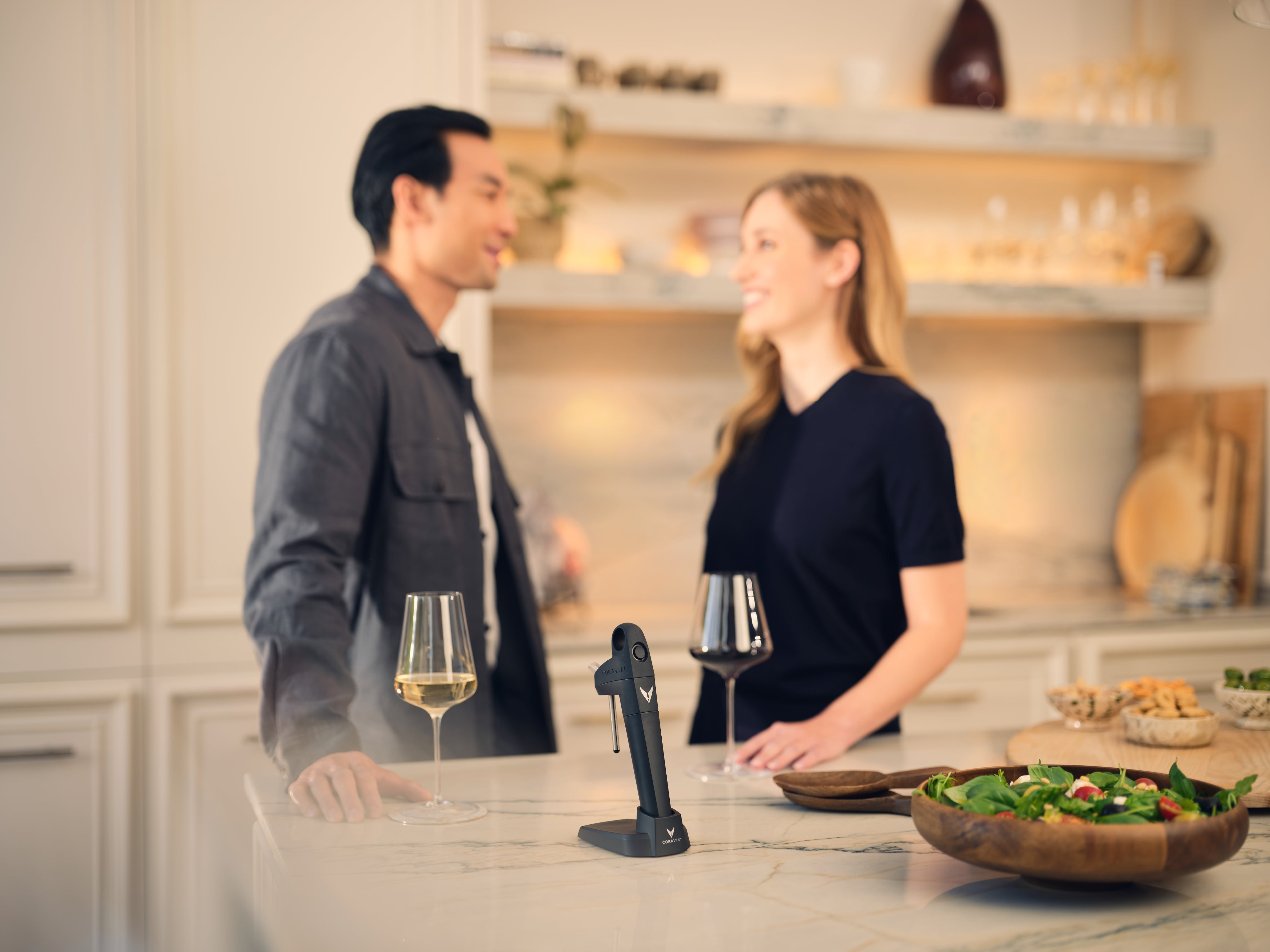 Two people in a kitchen with wine glasses , coravin pivot system and a salad.