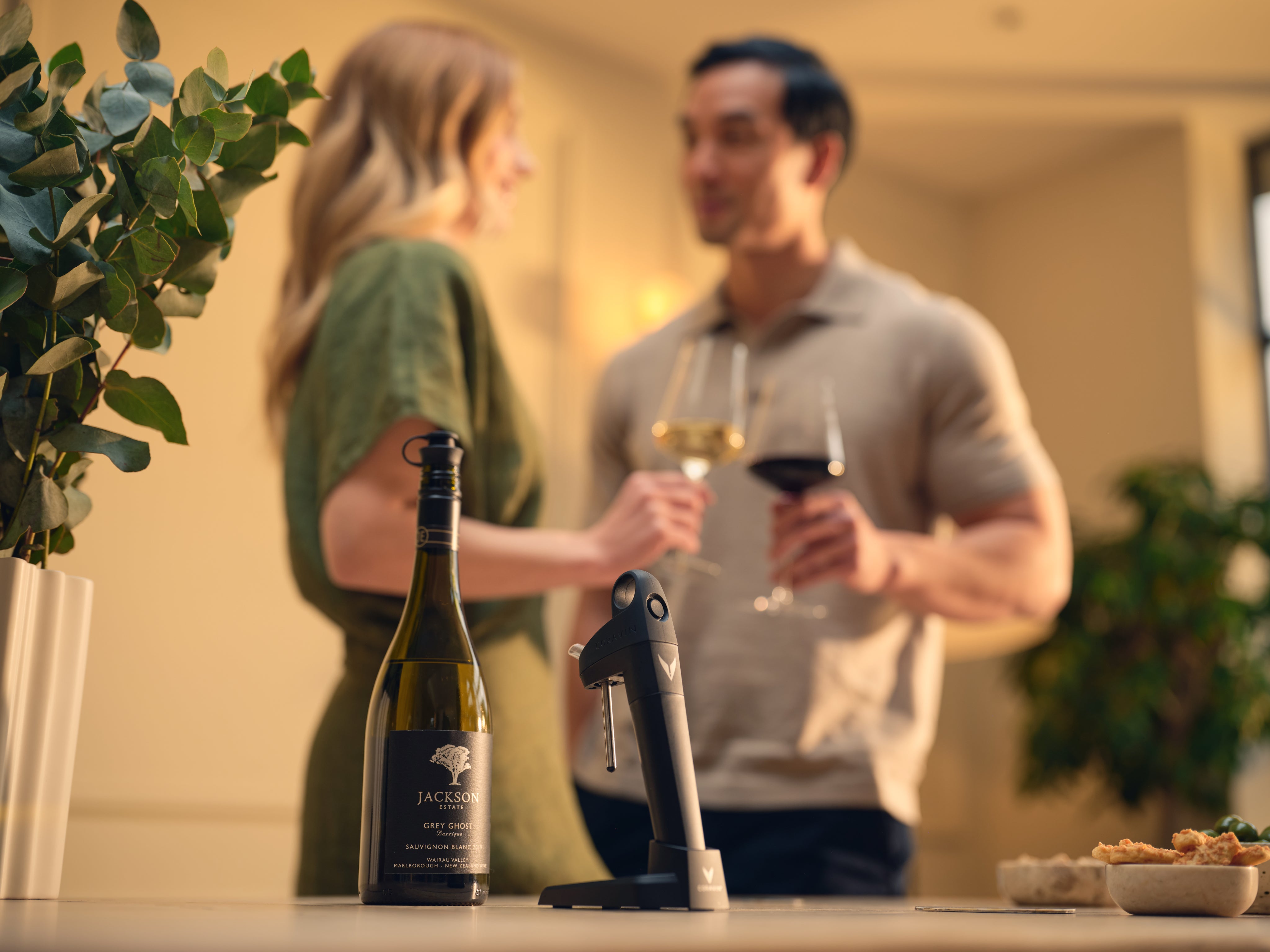 Two people standing in a kitchen with wine bottles and glasses, blurred background