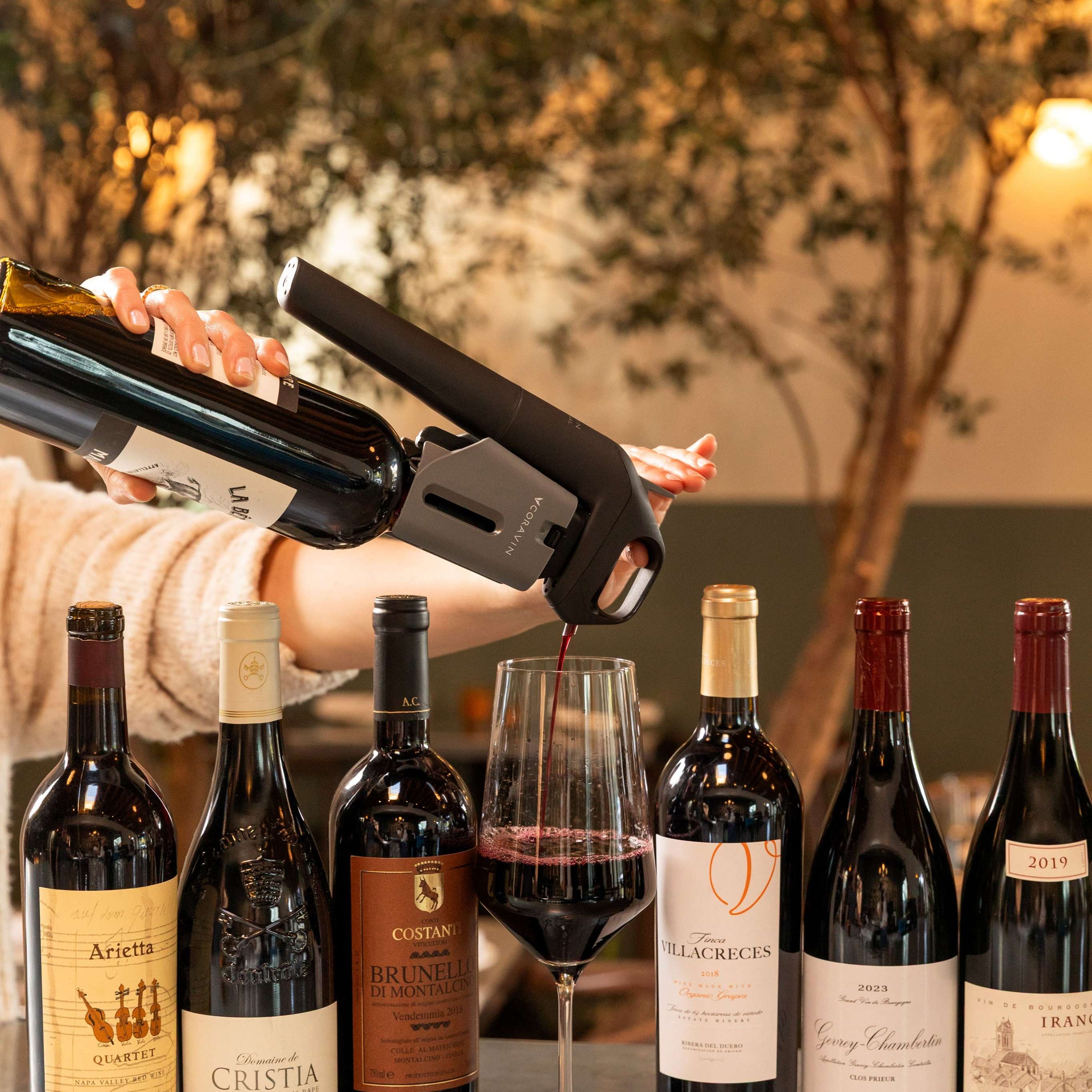 A Coravin Timeless system pours red wine into a glass, surrounded by premium still wine bottles.