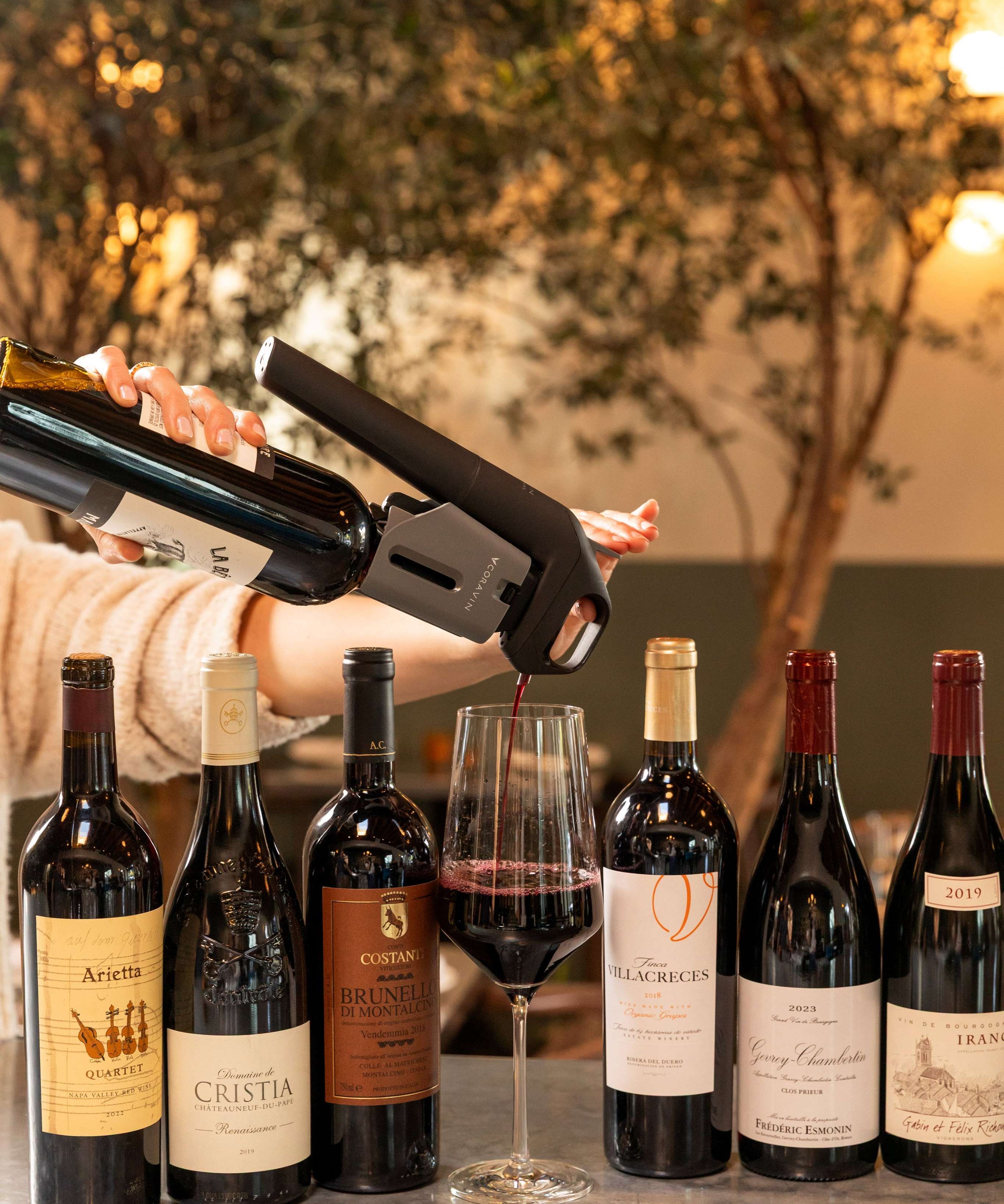 A Coravin Timeless system pours red wine into a glass, surrounded by premium still wine bottles.